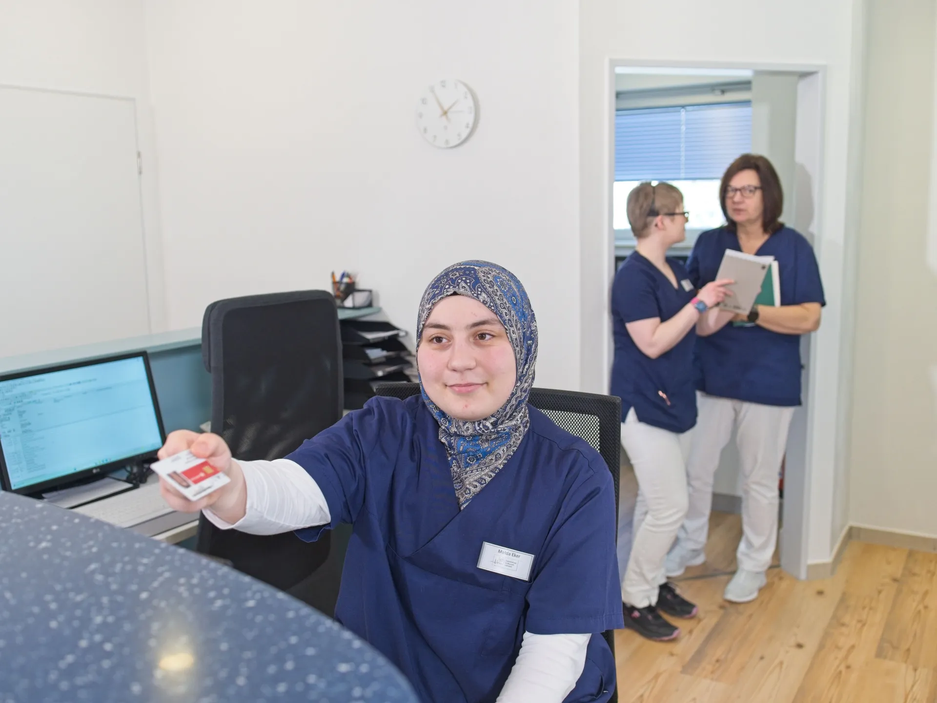 Eine medizinische Fachkraft in OP-Kleidung sitzt auf einem Wohnzimmersofa und misst den Blutdruck eines älteren Patienten mit einem digitalen Blutdruckmessgerät.